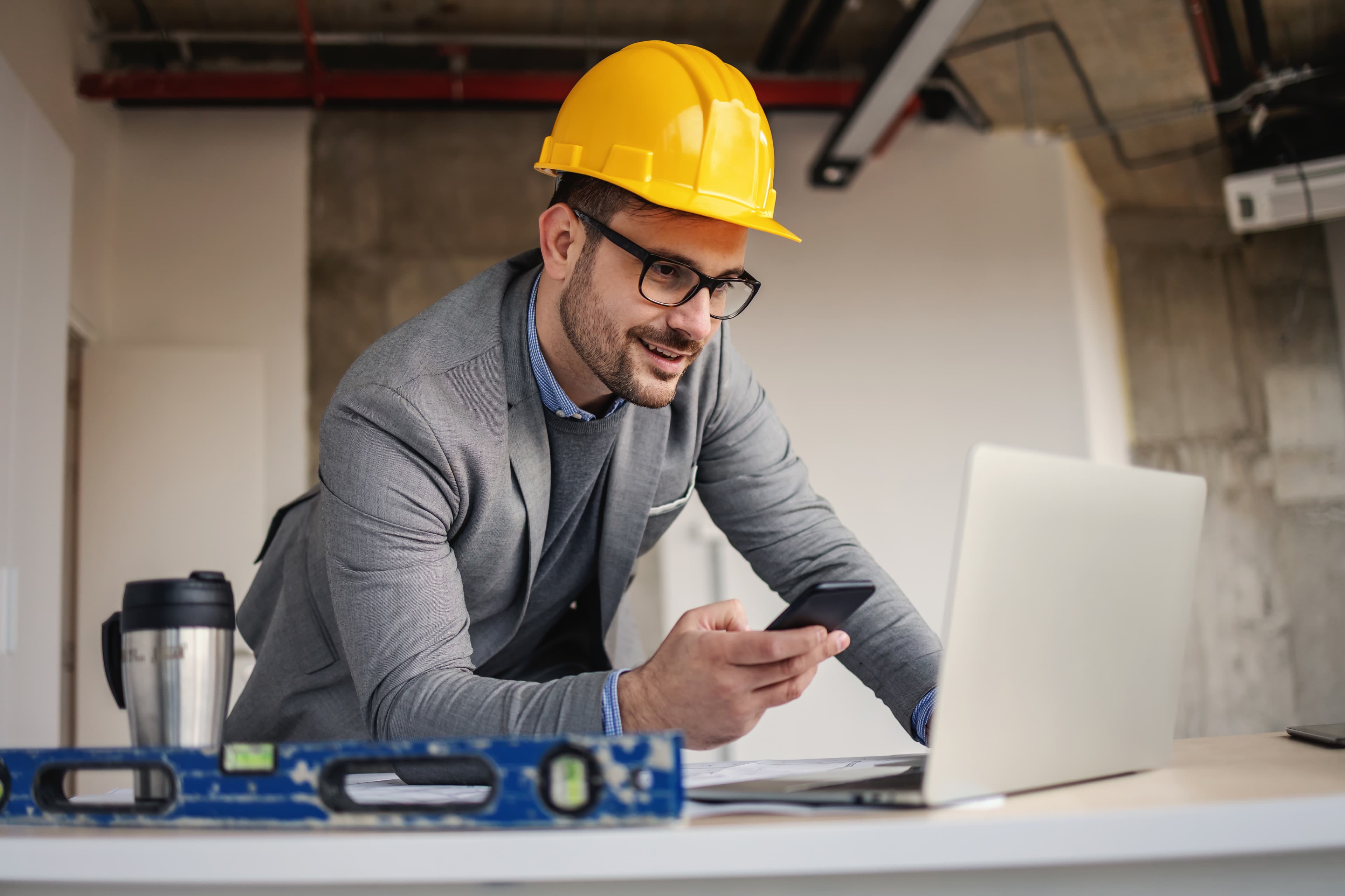 A contractor in a hard hat using a laptop and phone at a job site desk, accessing Pro Contractor Services at Aurora Home Hardware Building Centre.